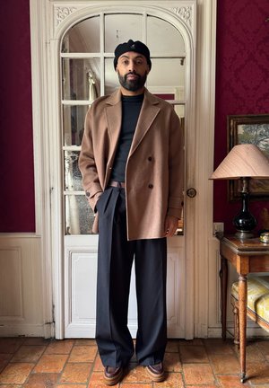 Rjav volnen plašč, črn prilagojen zgornji del, široke črne hlače, rjavi čevlji. Ozadje z vintage svetilko in leseno mizo. Rdeča stenska dekoracija.