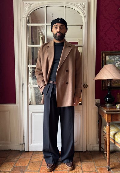 Abrigo de lana marrón, blusa negra ajustada, pantalones anchos negros, zapatos marrones. El fondo presenta una lámpara vintage y una mesa de madera. Decoración de pared roja.
