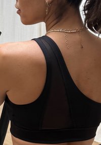 Černá sportovní podprsenka s hladkou texturou, širokými ramínky a síťovaným panelem na zádech. Vyznačuje se minimalistickým designem a trapezoidním tvarem.