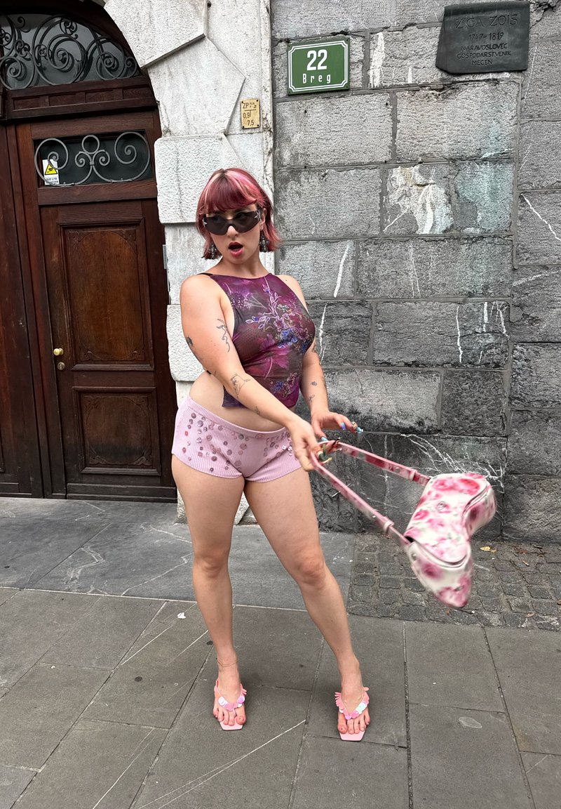 Personne aux cheveux roses, portant un haut violet et un short rose, faisant tourner un sac fleuri sur un trottoir près d'un mur en pierre et d'une porte en bois.