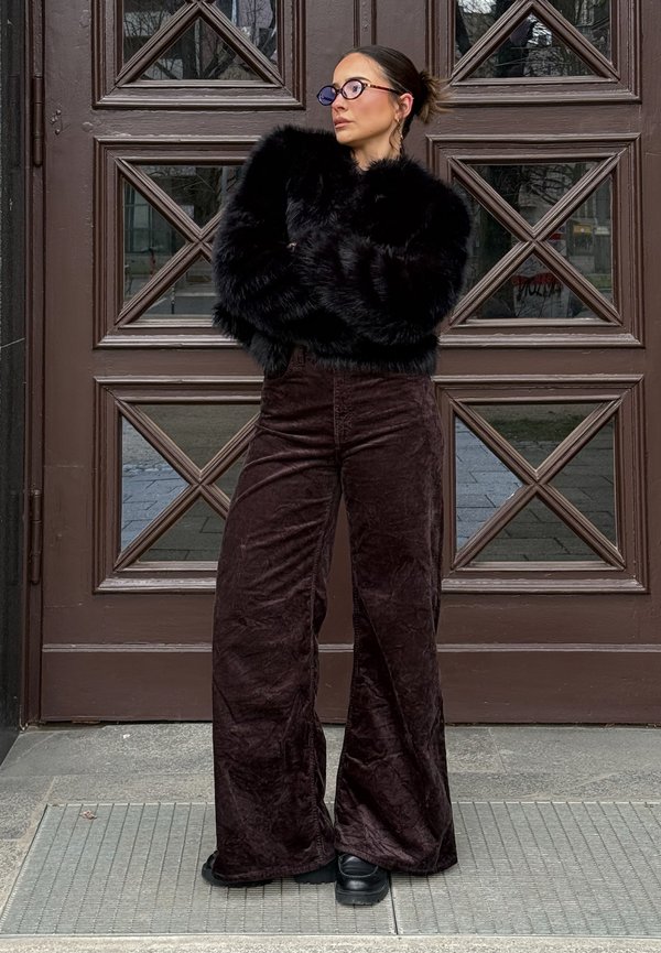 Mujer con el pelo oscuro recogido en un moño, lleva gafas negras, una chaqueta negra de piel sintética, pantalones anchos de terciopelo marrón y zapatos negros, está de pie con los brazos cruzados junto a una puerta de madera.