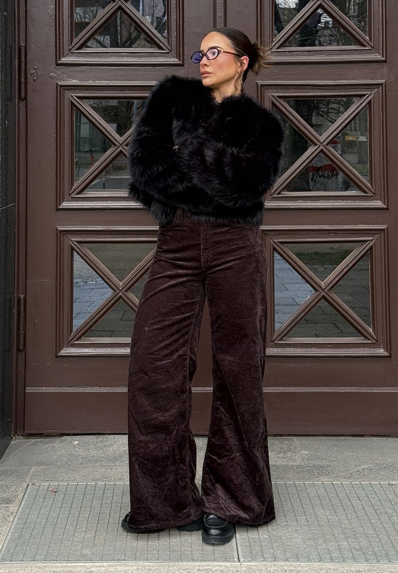 Γυναίκα με σκούρα μαλλιά σε κότσο, φοράει μαύρα γυαλιά, μαύρο μπουφάν από ψεύτικη γούνα, φαρδιά καφέ βελούδινα παντελόνια και μαύρα παπούτσια, στέκεται με τα χέρια σταυρωμένα κοντά σε μια ξύλινη πόρτα.