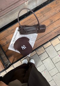 Brun læder håndtaske med lynlås, brun kasket med hvidt logo, og hvide sneakers på en træbænk med en avis på fliseoverfladen.