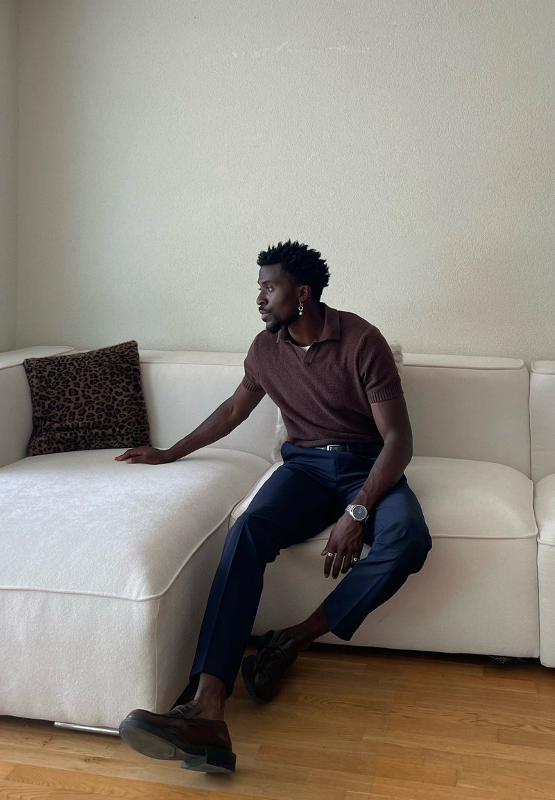 Uomo seduto che indossa una camicia marrone a maniche corte e pantaloni blu navy su un divano bianco, con un braccio poggiato su un cuscino con stampa leopardata.