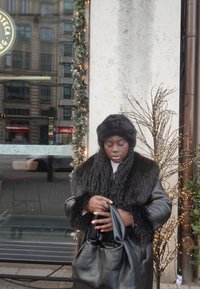 Zwarte nepbontjas met een getextureerde kraag, gecombineerd met een bijpassende bontmuts. Houdt een zwarte totebag vast, staand tegen een feestelijke achtergrond.