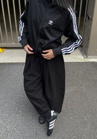Nero giacca a zip Adidas con strisce bianche sulle maniche, pantaloni larghi neri e scarpe da calcio nere con strisce bianche. Anelli visibili sulle dita.