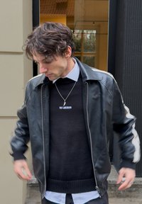 Jaqueta bombardeiro de couro preta com riscas brancas nas mangas, sobre um suéter de malha preto, vestida com uma camisa branca de colarinho.