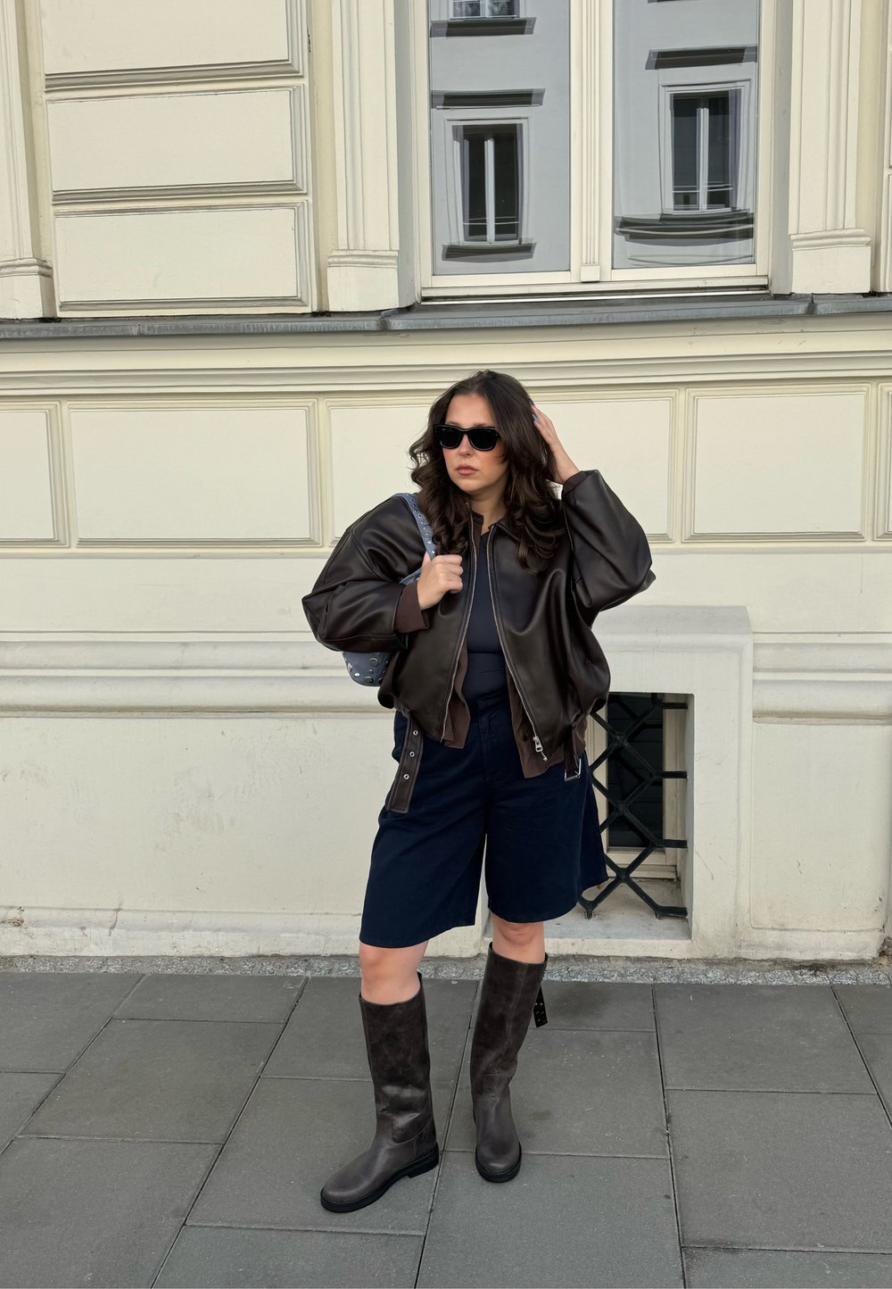Hnědá kožená bomber bunda, černé šortky, tmavě hnědé kozačky po kolena a černé sluneční brýle. Hladká textura s uvolněnou siluetou.