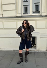 Hnědá kožená bomber bunda, černé šortky, tmavě hnědé kozačky po kolena a černé sluneční brýle. Hladká textura s uvolněnou siluetou.