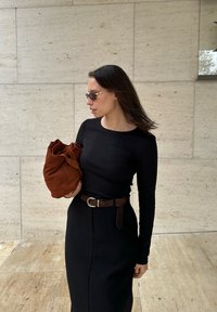 Melna garām piedurknēm blūze, augstas jostas melna svārku, brūna zamša soma un tumši saulesbrilles ar zelta akcentiem, novietotas uz akmens fona.