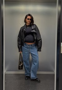 Veste en cuir noire, haut moulant noir, jean à jambes larges bleu clair et chaussures noires. Tient un sac à main foncé, avec des lunettes de soleil.