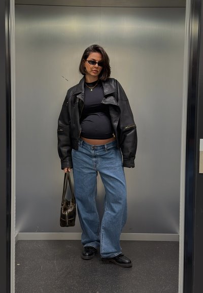 Chaqueta de cuero negra, camiseta negra ajustada, vaqueros anchos de color azul claro y zapatos negros. Sosteniendo un bolso oscuro, con gafas de sol.
