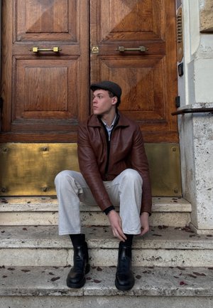 Chaqueta de cuero marrón, camisa azul plata, pantalones gris claro, botas negras de tobillo, sentado en escalones de piedra frente a puertas de madera con acentos de bronce.