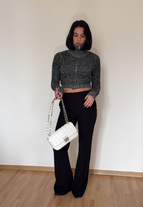 Pantaloni neri a vita alta e un maglione grigio corto con zip. Tiene una borsa bianca trapuntata con una tracolla in catena argentata. Sfondo in legno.