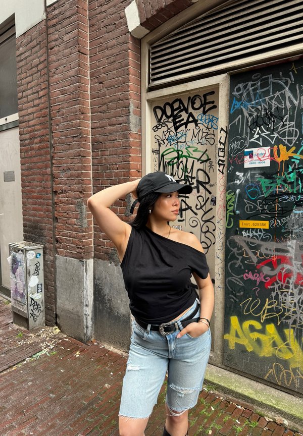 Svart av-shoulder topp, lyseblå slitne denimshorts, svart belte med sølvspenne, og svart caps. Murvegg og graffiti-bakgrunn.