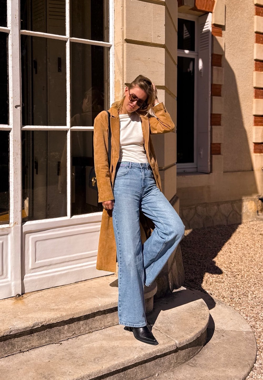 Korte tan suède lange jas over een witte top, lichtblauwe wijde jeans en zwarte puntige enkellaarsjes. Staande op stenen trappen bij een raam.