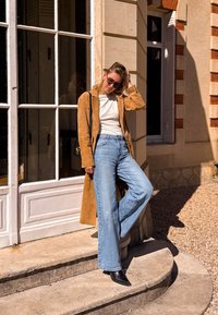 Korte tan suède lange jas over een witte top, lichtblauwe wijde jeans en zwarte puntige enkellaarsjes. Staande op stenen trappen bij een raam.