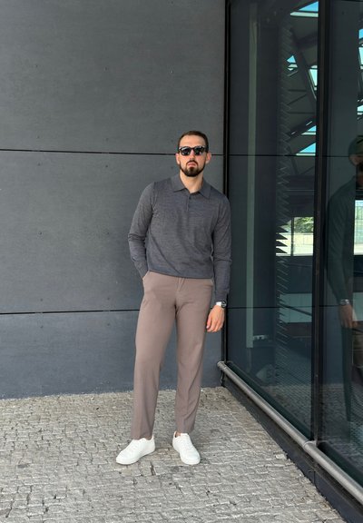 Camisa polo gris de manga larga, pantalones de vestir marrón claro, zapatillas blancas y gafas de sol. De pie contra una pared de cristal con una superficie texturizada.