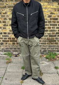 Osoba u crnoj jakni na patentni zatvarač, zelenim cargo hlačama i crnim cipelama stoji na pločniku ispred zidane zid.