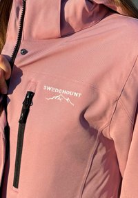 Helle rosa Outdoorjacke mit strukturiertem Stoff. Verfügt über eine Zipper-Tasche und das "SWEDEMOUNT"-Logo in Weiß, zusammen mit Bergen-Grafiken.