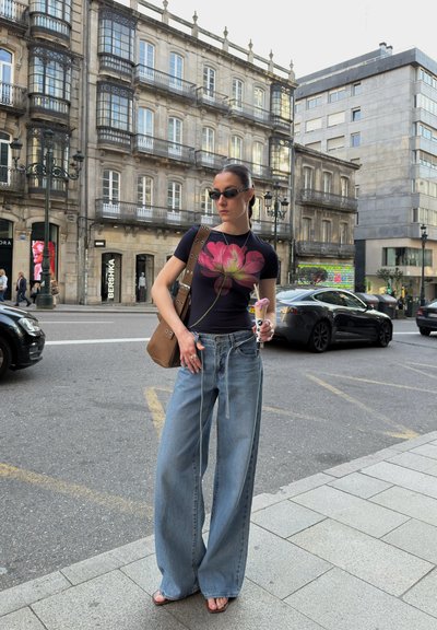 Camiseta negra con una gran flor de loto rosa, jeans de pierna ancha de color azul claro, bolso de hombro marrón, gafas de sol, sosteniendo un cono de helado.