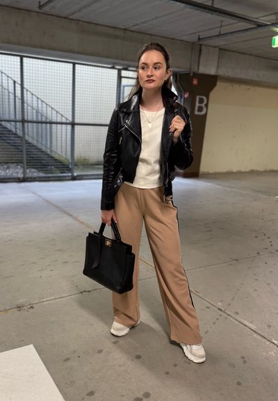 Chaqueta de cuero negra sobre una blusa blanca, combinada con pantalones de pierna ancha beige con rayas negras. Sosteniendo un bolso negro y usando zapatillas blancas.