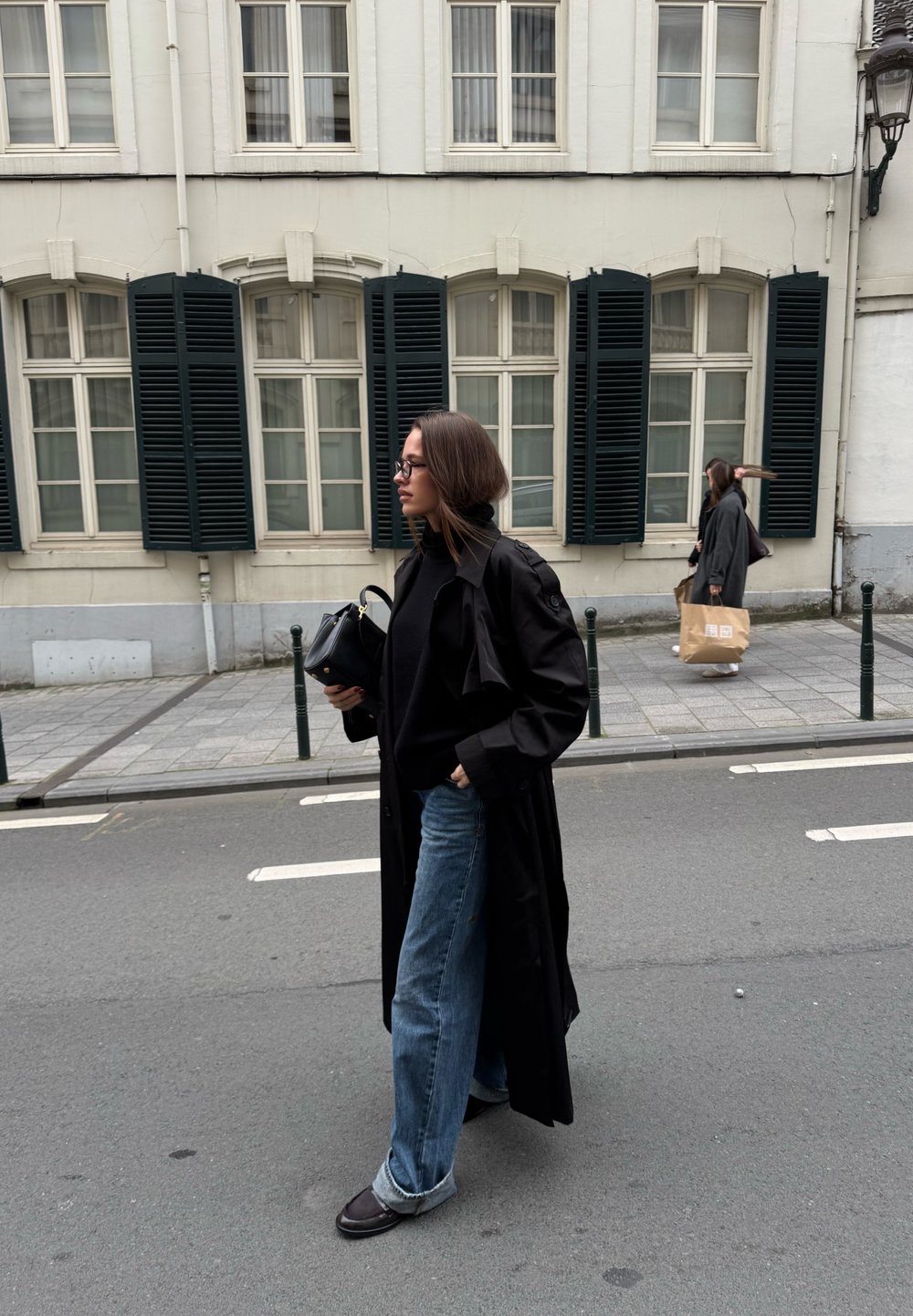 Čierny trenčkot cez čierny sveter, voľné modré džínsy a tmavá obuv. V pozadí je budova s bielymi stenami a tmavými žalúziami.