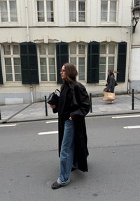 Čierny trenčkot cez čierny sveter, voľné modré džínsy a tmavá obuv. V pozadí je budova s bielymi stenami a tmavými žalúziami.