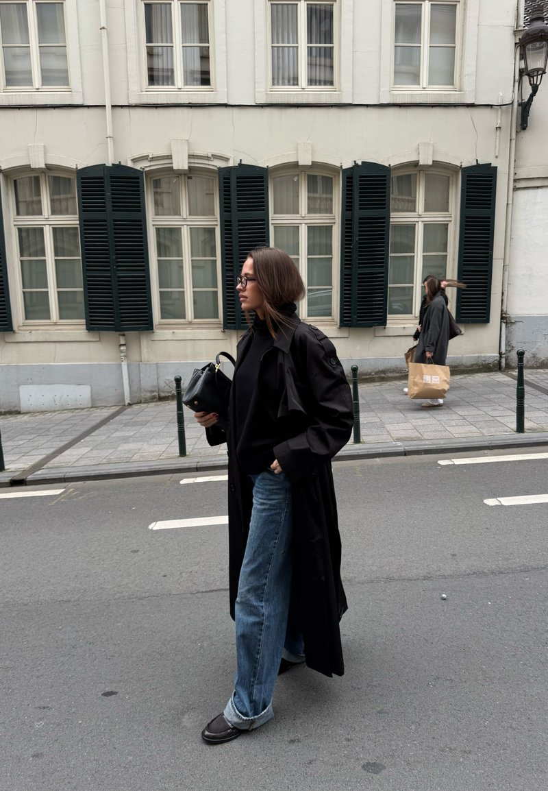 Black trench coat over a black sweater, loose blue jeans, and dark shoes. The background features a building with white walls and dark shutters.