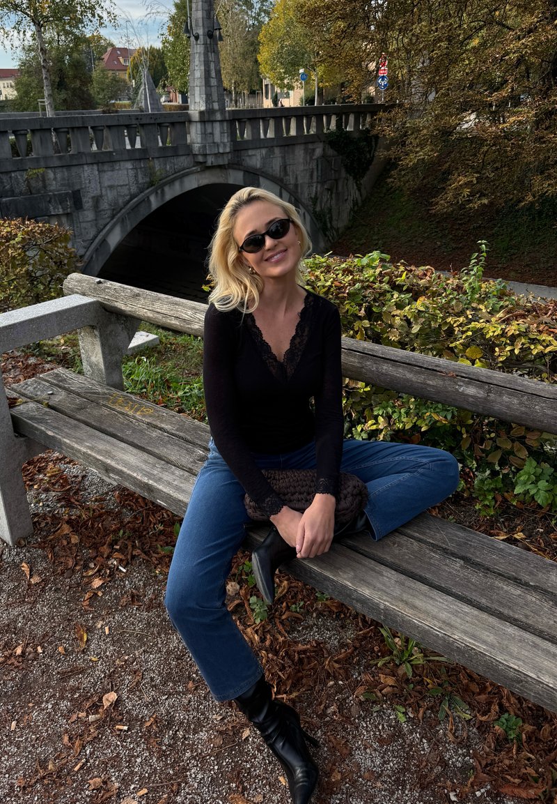 Une femme portant des lunettes de soleil est assise sur un banc en bois près d’un vieux pont de pierre, entourée de feuillage automnal.