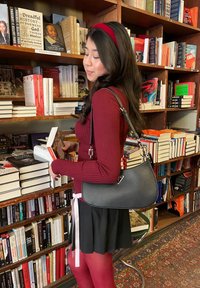 Bolso de hombro negro con forma curvada y herrajes plateados, combinado con un suéter de canalé burdeos y una falda negra, en un entorno de librería.