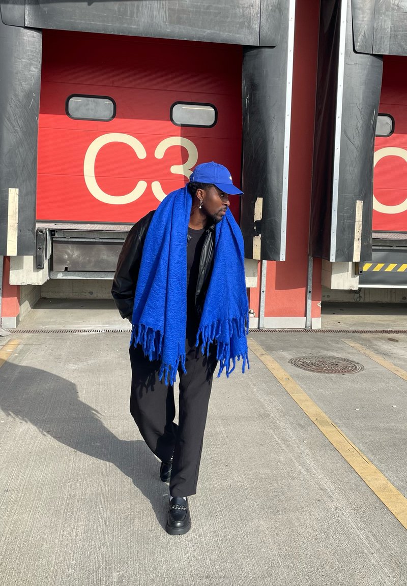 Man wearing a large blue scarf and blue cap walking in front of a red loading dock door marked "C3" in an industrial area.