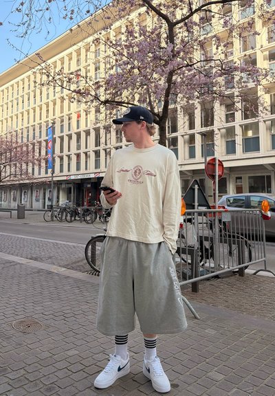 Joven con ropa casual holgada y gorra, de pie en la acera de la ciudad, sosteniendo un teléfono, con un árbol en flor y un edificio moderno al fondo.