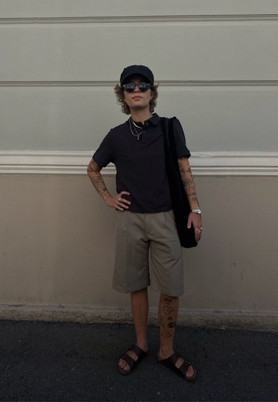 Persona vistiendo una camiseta polo negra, pantalones cortos beige, sandalias oscuras y gafas de sol. Sosteniendo un bolso negro, mostrando tatuajes intrincados en los brazos.