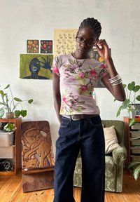 Camisa justa de mangas curtas com base rosa e padrão floral tropical. Combinada com jeans de perna larga em azul escuro. Cadeira de elefante esculpida em madeira ao fundo.