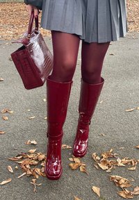 Βουργουνδίας παλτό δερμάτινα μποτάκια μέχρι το γόνατο με γυαλιστερό φινίρισμα και αξεσουάρ με πόρπες, συνδυασμένα με γκρι πτυχώσεις φούστα και τσάντα που ταιριάζει.