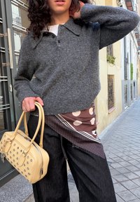 Personne en plein air portant un pull gris, une écharpe à motifs, un pantalon noir, tenant un sac à main beige clouté, debout sur une rue pavée.