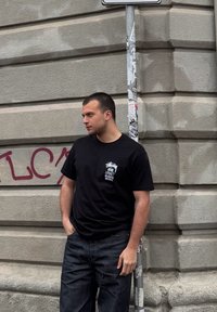 Mlad moški s kratkimi lasmi, oblečen v črn majico z grafiko in temne kavbojke, se opira ob teksturirano betonsko steno z grafiti in stebrom pokritim s nalepkami.