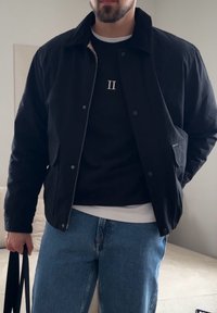 Homme portant une veste noire sur un pull noir avec "II" sur la poitrine, un jean bleu clair, tenant une sangle de sac noire à l'intérieur.