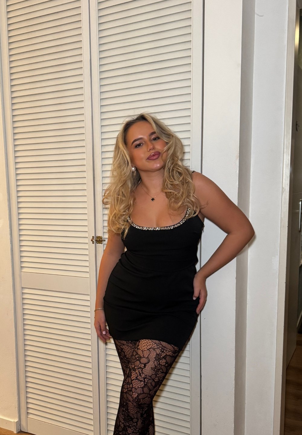 Femme blonde en robe noire avec un col orné de perles posant devant des portes blanches à persiennes, portant des collants en dentelle noire et des boucles d'oreilles en perles.
