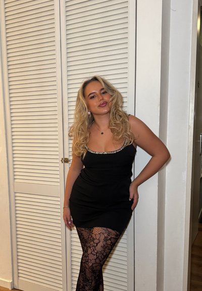 Mujer rubia con un vestido negro de cuello con cuentas que posa frente a puertas blancas con persianas, lleva medias negras de encaje y pendientes de perlas.