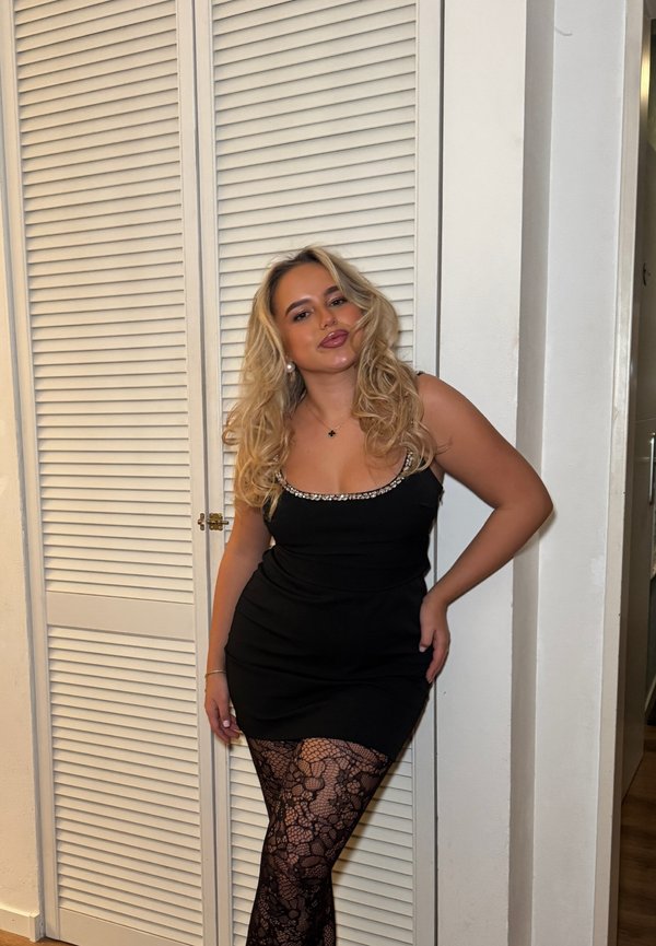 Mujer rubia con un vestido negro de cuello con cuentas que posa frente a puertas blancas con persianas, lleva medias negras de encaje y pendientes de perlas.