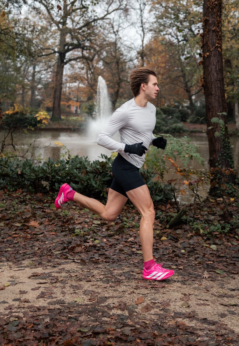 A runner in a light grey long-sleeve top and black shorts, wearing pink trainers, jogs along a leaf-covered path beside a pond.