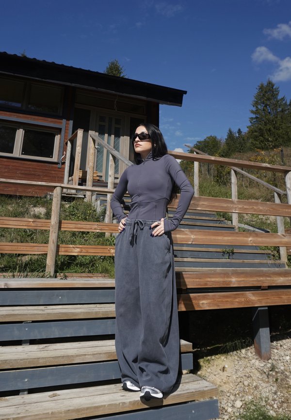 Mujer con el pelo largo y oscuro y gafas de sol, de pie en escaleras de madera fuera de una cabaña, vestida con una camiseta gris ajustada y pantalones anchos grises bajo un cielo azul despejado.