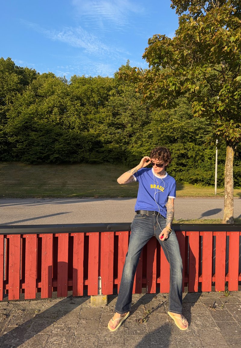 Jeune personne avec des tatouages portant des lunettes de soleil, un t-shirt bleu Brésil, un jean et des tongs, debout près d'une clôture en bois rouge près des arbres et de la route.