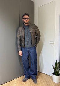 Homme portant des lunettes de soleil noires, un pull gris, une veste en cuir marron, un jean large bleu et des bottes grises, debout à l'intérieur près d'une porte blanche et d'une plante.