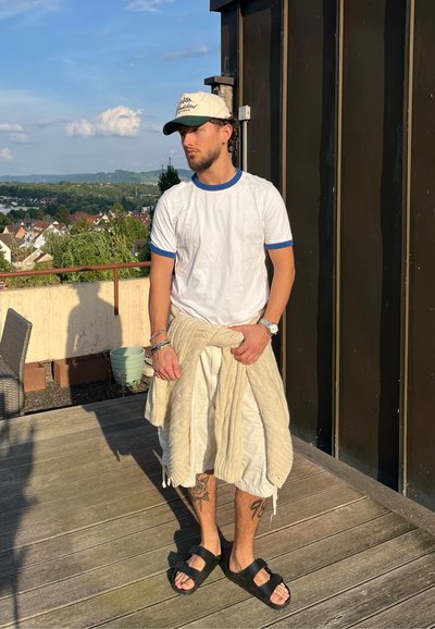 Joven con camiseta blanca, jersey beige atado a la cintura, pantalones cortos, sandalias negras y gorra beige, de pie en una terraza de madera con vistas al pueblo detrás.