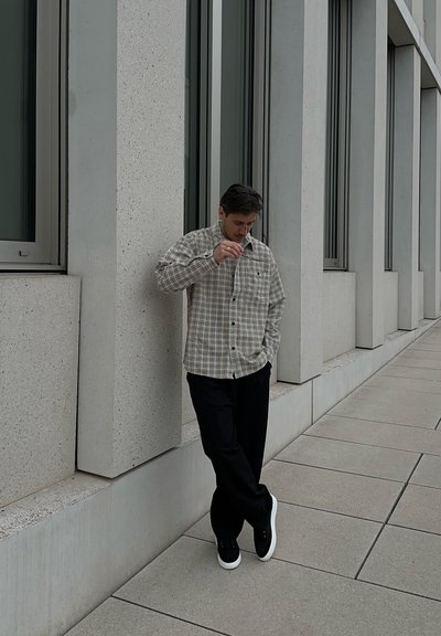 Hombre con camisa de cuadros y pantalones negros apoyado contra la pared de un edificio moderno con grandes ventanas, mirando hacia abajo con una mano cerca de su rostro.