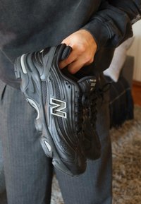 Käsi hoiab paari musti New Balance 1906 tossusid, millel on paelad, seljas on hallid püksid ja tume dressipluus siseruumides.