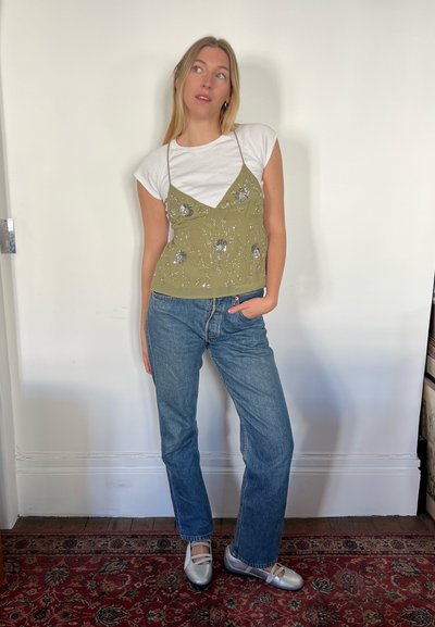 Camiseta de tirantes de un verde oliva claro con bordados florales, sobre una camiseta blanca, combinada con unos vaqueros rectos de un azul claro y bailarinas plateadas.